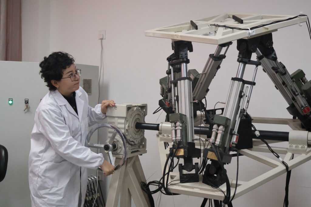Una mujer con bata blanca de laboratorio y gafas opera un volante manual de calibración conectado a una estructura metálica e hidráulica de alta tecnología.