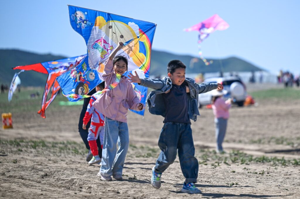 Dos niños sonrientes corren al aire libre en un terreno despejado sosteniendo cometas o barriletes coloridos, con montañas lejanas bajo un cielo claro de fondo.