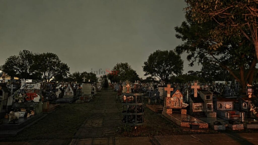 Foto nocturna del pasillo central del Cementerio de Florencio Varela con cruces iluminadas y una figura misteriosa al fondo.