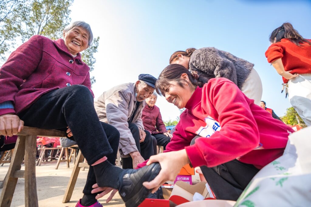 Una voluntaria o asistente coloca calzado nuevo a una mujer mayor sonriente durante una actividad comunitaria al aire libre en China.