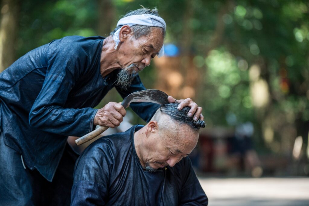 Hombre mayor de una minoría étnica china realizando un corte de cabello tradicional con una hoz curva a otro anciano en un entorno natural.