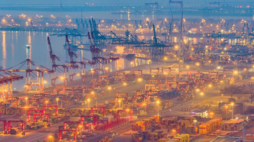 Vista panorámica nocturna de un concurrido puerto de carga chino con grúas operativas y miles de contenedores.   