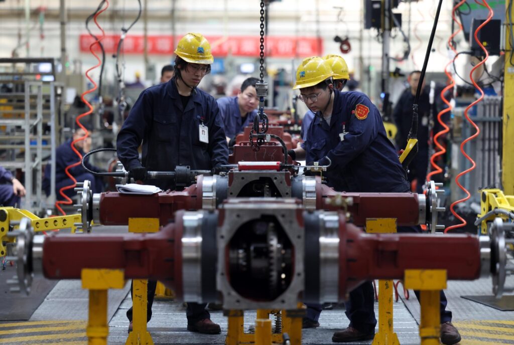 Dos trabajadores de industria pesada con cascos amarillos ensamblando componentes mecánicos en una línea de montaje en China.