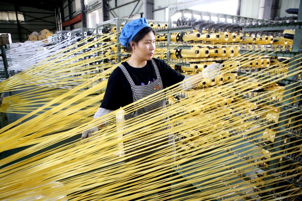 Trabajadora en una fábrica textil en China supervisando maquinaria con múltiples hilos amarillos extendidos.